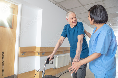 Asian female doctor reassuring mature elderly man with walker. Man and woman smiling happy in the hospital aisle. Retirement community concept