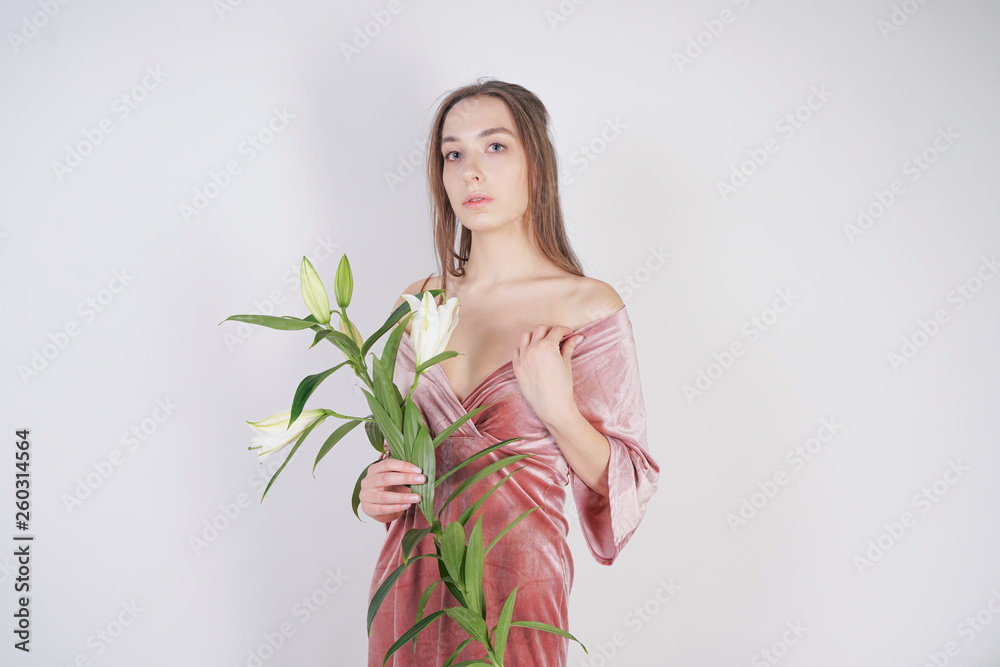 shy caucasian girl with big eyes stands in a velvet pink dress and holds a white Lily in her hand as a symbol of innocence and purity. young woman posing on white studio background. shy caucasian girl with big eyes stands in a velvet pink dress and holds a white Lily in her hand as a symbol of innocence and purity. young woman posing on white studio background.