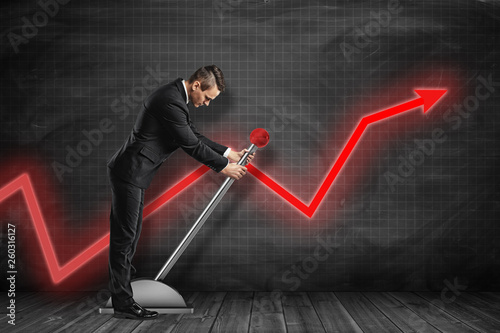 Side view of businessman looking down and pulling big lever near black graph ruled wall with red graph arrow on it.