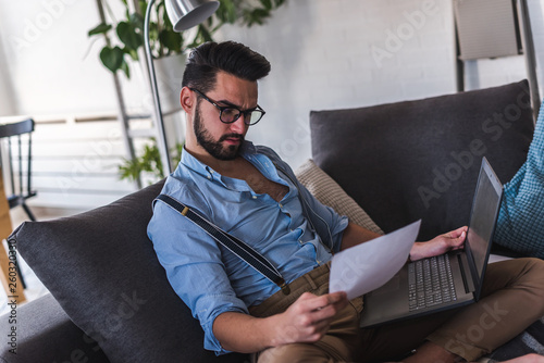 Young bearded businessman is sitting on sofa working with laptop computer. Freelancer, entrepreneur works at home. photo