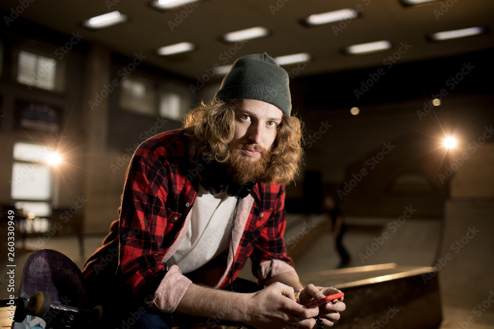 Waist up portrait of bearded man looking at camera while sitting on ramp in extreme park, shot with flash