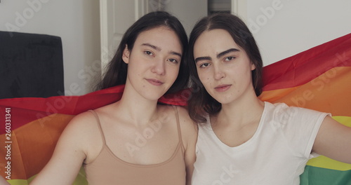 Portrait of lesbian couple with LGBT flag looking into the camera. Two happy girlfriends pose with rainbow flag at home.