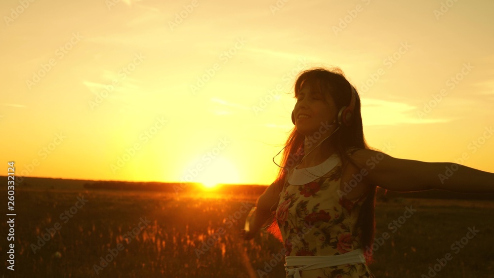 young girl in headphones and with a smartphone whirl in flight under the rays of a warm sunset. Slow motion. Happy girl listening to music and dancing in the rays of beautiful sunset in the park.