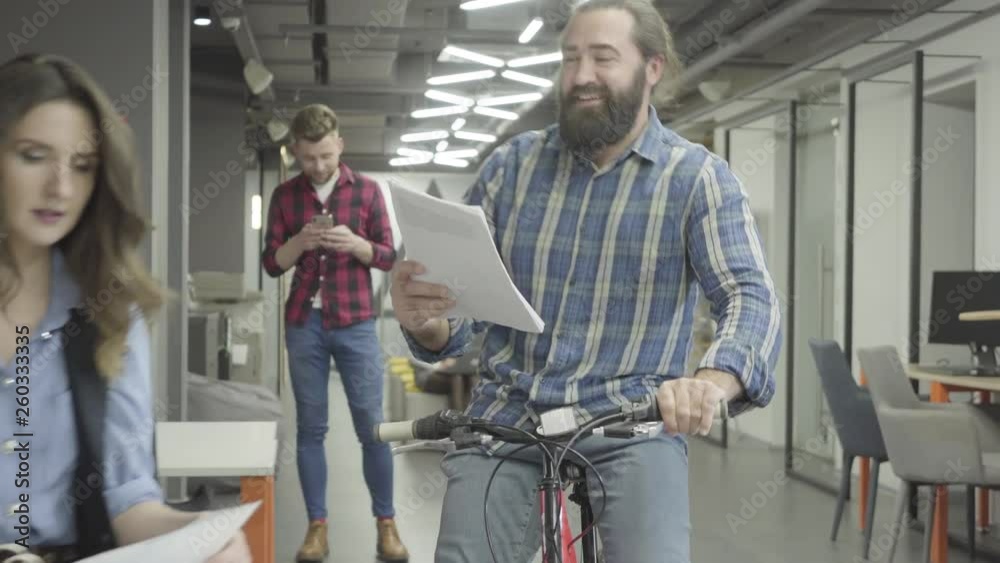 Bearded smiling man rides his bike with papers in hand and shows documents to the lady in the office. Pretty woman and bearded man communicate in the office, discussing new document, they are excited Bearded smiling man rides his bike with papers in hand and shows documents to the lady in the office. Pretty woman and bearded man communicate in the office, discussing new document, they are excited