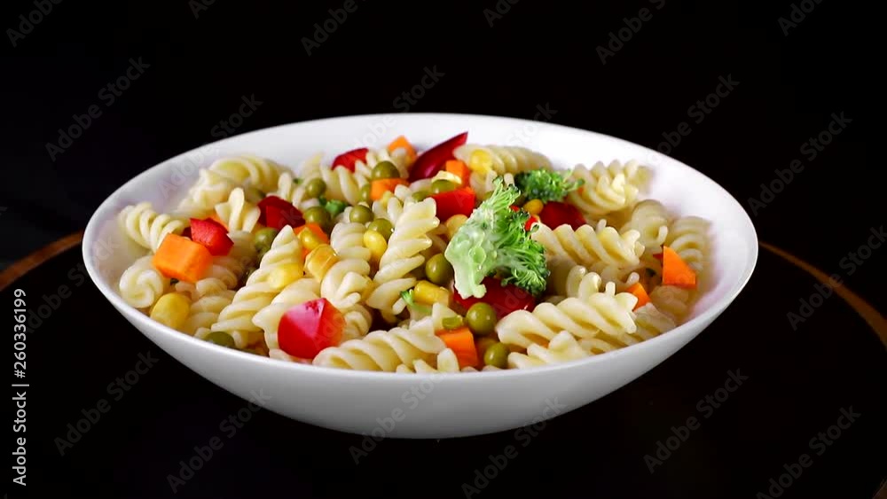Spiral pasta with vegetables on a plate revolve on a black background. Green cauliflower falls on a plate Spiral pasta with vegetables on a plate revolve on a black background. Green cauliflower falls on a plate