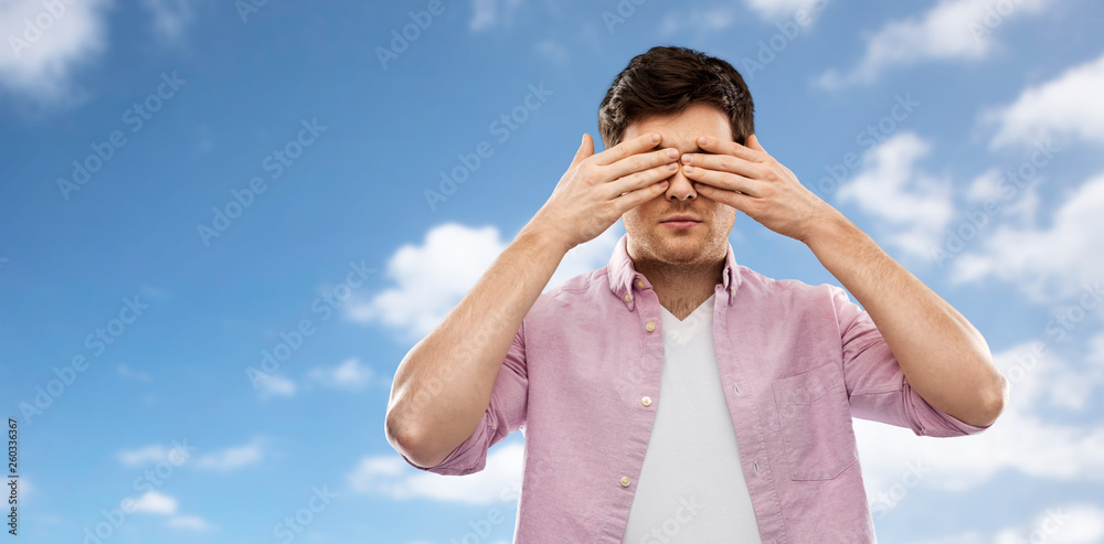 Fototapeta premium vision and people concept - man closing his eyes by hands over blue sky and clouds background