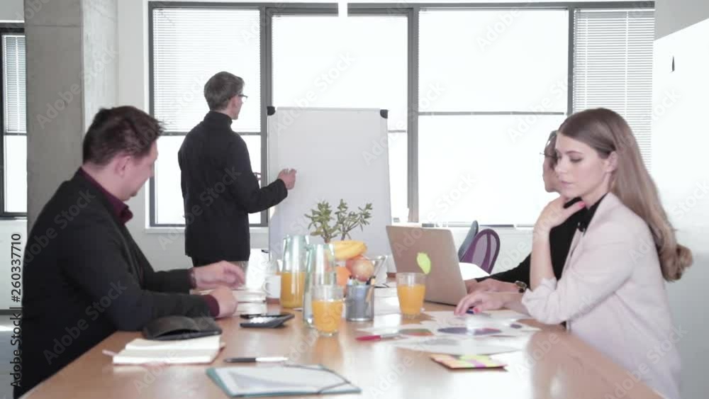 Candid moment, middle age male manager presents new project plan to colleagues at meeting. Mature businessman explaining ideas on flipchart to coworkers in office. Candid moment, middle age male manager presents new project plan to colleagues at meeting. Mature businessman explaining ideas on flipchart to coworkers in office.