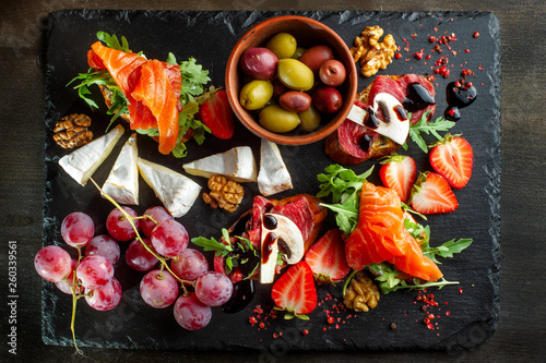 Snacks laid out on black slate, cheese, grapes, sandwiches, strawberries, nuts.