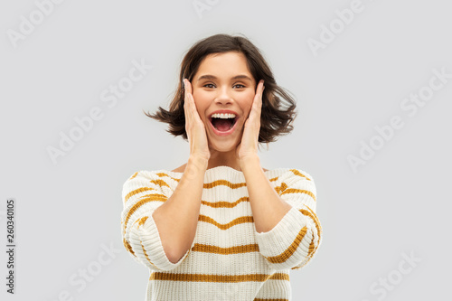 emotion, expression and people concept - happy impressed young woman in striped pullover holding to her face over grey background