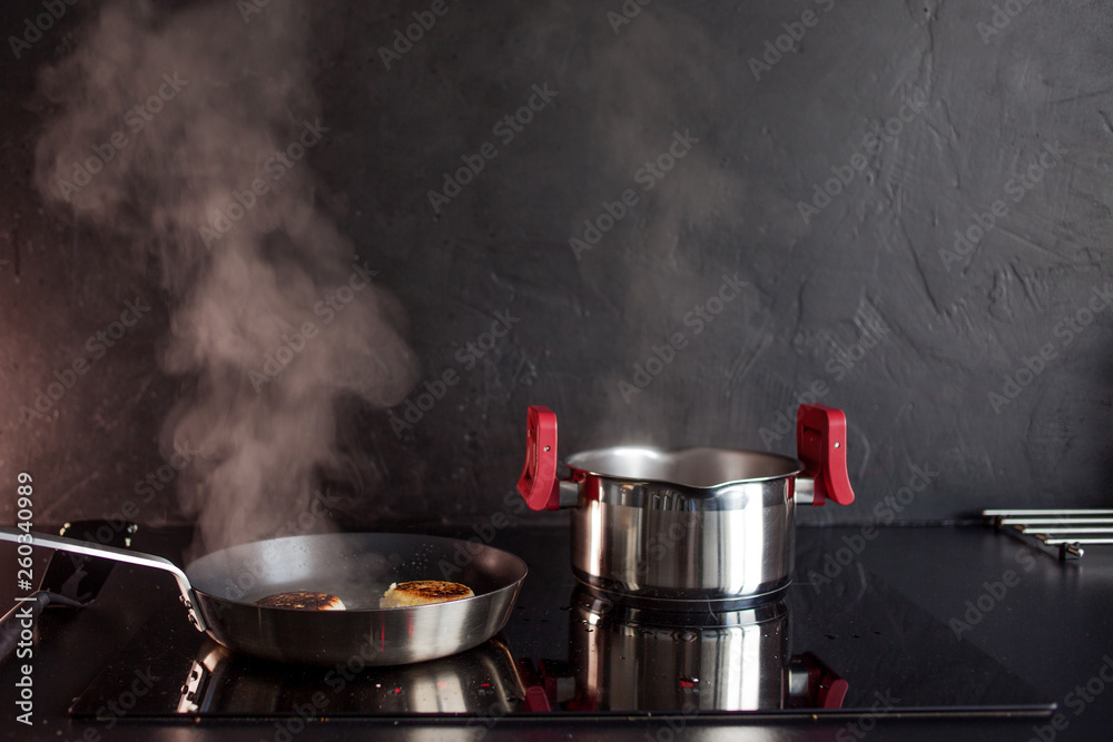 Frying pan and cooking pot on induction hob, steam rises. Black textured kitchen