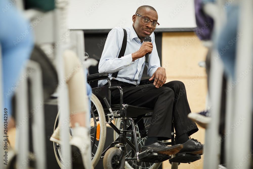 Serious confident motivational disabled mature African-American speaker in glasses sitting in wheelchair and looking at audience while saying speech into microphone at conference - obrazy, fototapety, plakaty 