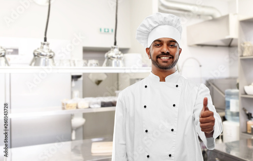 cooking, profession and people concept - happy male indian chef in toque showing thumbs up over restaurant kitchen background