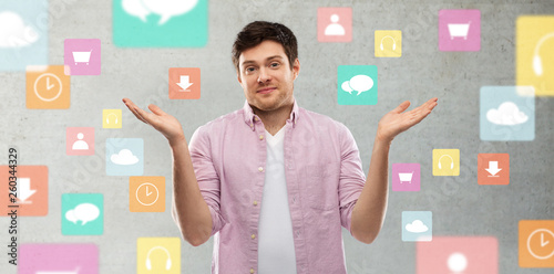 people, technology and expression concept - young man shrugging over app icons on grey background