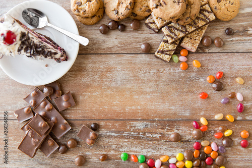 junk food, sweets and unhealthy eating concept - close up of chocolate, drop candies and piece of cake and cookies on wooden table