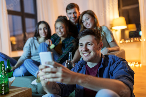 friendship, technology and entertainment concept - happy friends with smartphone, snacks and non-alcoholic drinks hanging out at home in evening