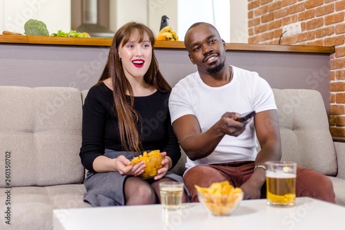 Ethnic family sitting on the couch in the living room watching TV drinking beer eating chips