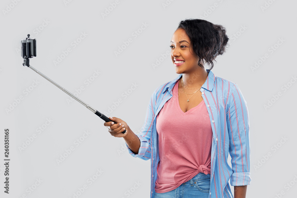 technology and people concept - happy african american woman taking picture by smartphone on selfie stick over grey background technology and people concept - happy african american woman taking picture by smartphone on selfie stick over grey background