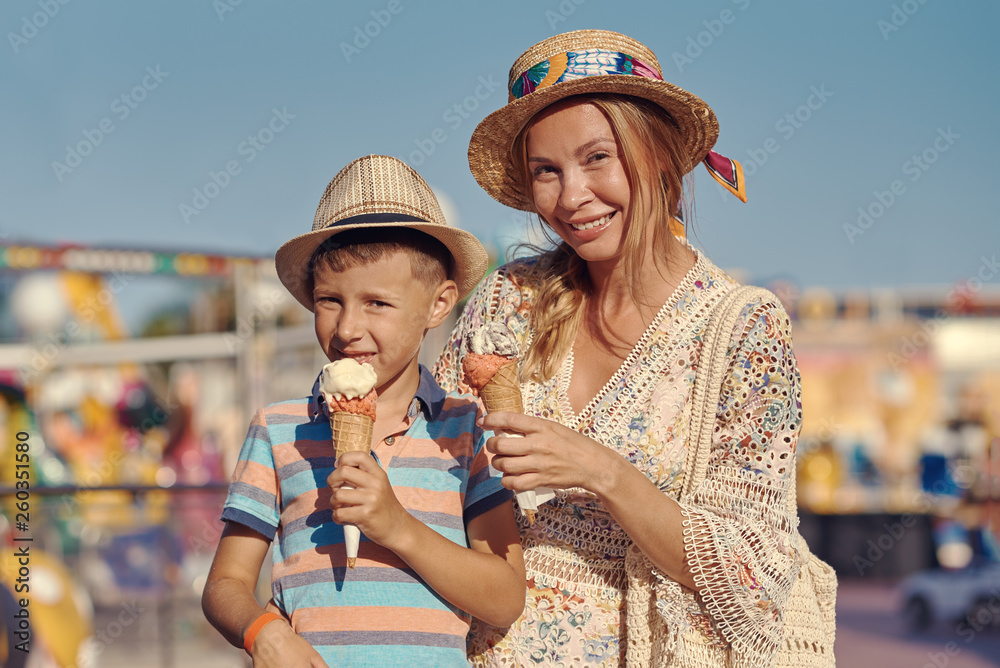 Cute European boy and his mother are walking around the amusement park and eating tasty ice-cream.
