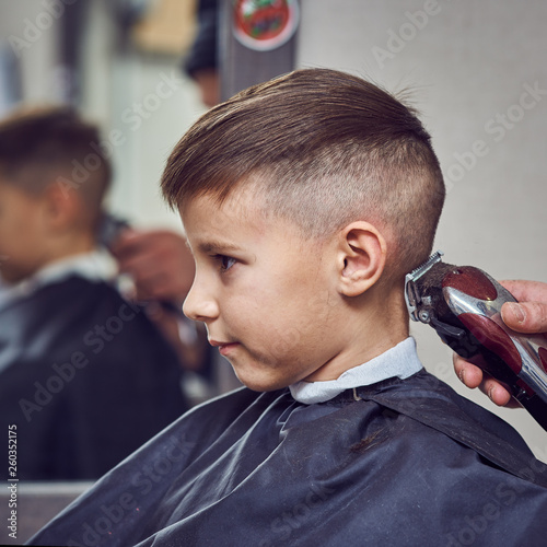 Barber making a haircut to a cute European boy using cutting machine.
