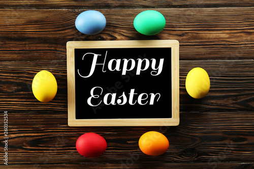 Blackboard with Happy Easter text, different colorful boiled paintd eggs. Greeting card concept with traditional holiday attributes. Wooden background, close up, top view, copy space, flat lay.