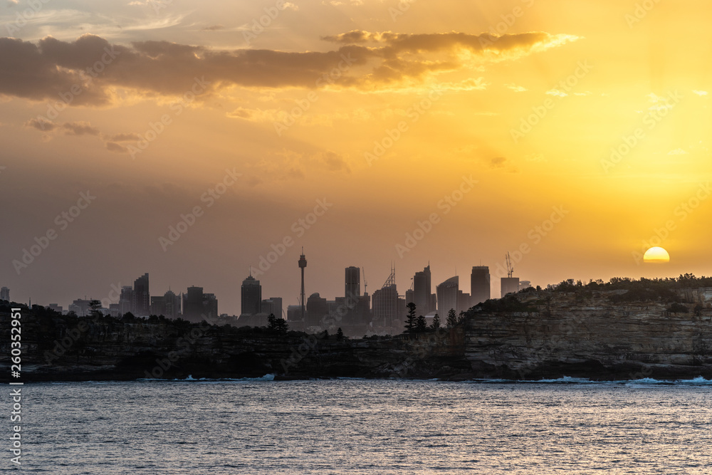 Sydney, Australia - February 12, 2019: Sunset over city skyline seen from Tasman Sea. Shoreline rocky cliffs. Yellow brown sky, sun rays. 4 of 5.