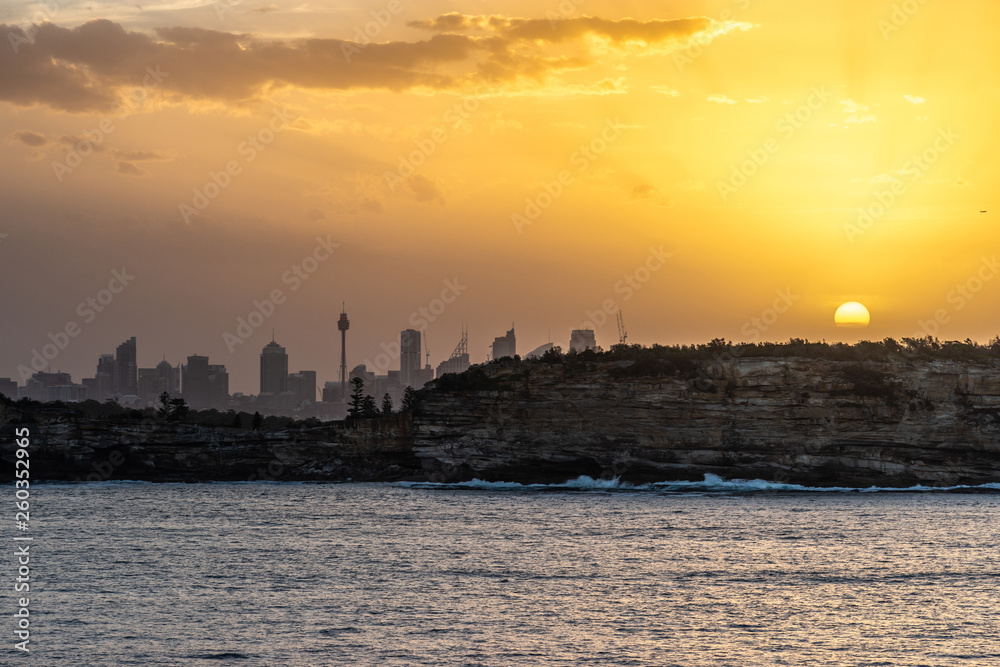 Fototapeta premium Sydney, Australia - February 12, 2019: Sunset over city skyline seen from Tasman Sea. Shoreline rocky cliffs. Yellow brown sky, sun rays. 3 of 5.