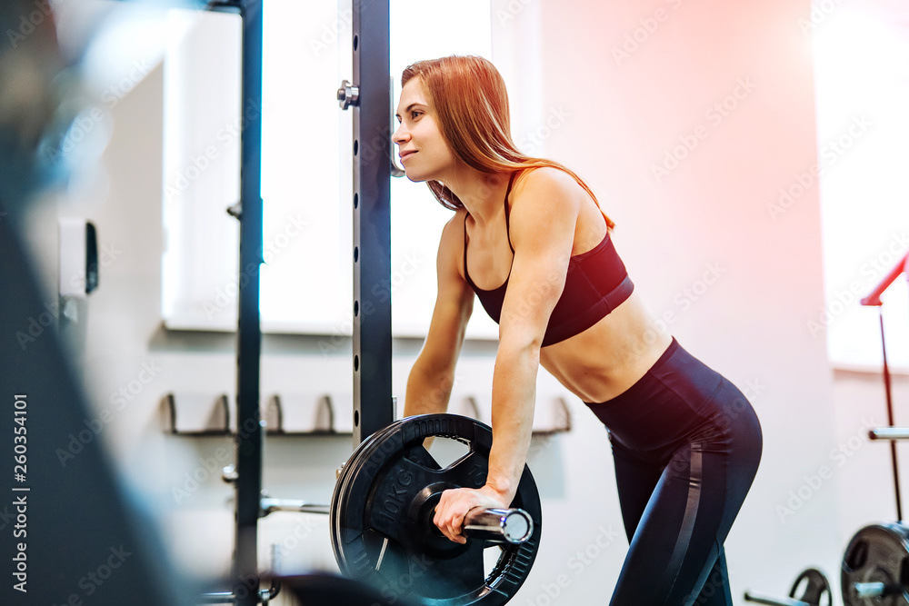 Beautiful happy young sportswoman in fitness clothes standing near the heavy barbell in the gym. Smiling female doing weight lifting workout at sports center background. Beautiful happy young sportswoman in fitness clothes standing near the heavy barbell in the gym. Smiling female doing weight lifting workout at sports center background.