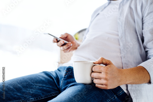 A midsection of man with smartphone and coffee sitting indoors, text messaging.