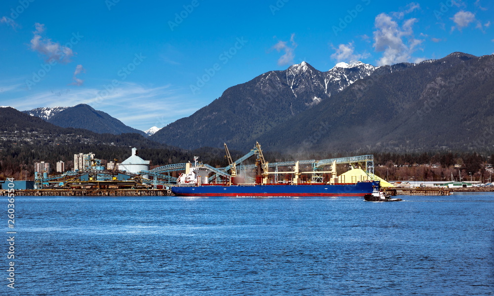 Terminal of North Vancouver port on a background of mountain scenery. The vessel is under load, the tug is towing another vessel. Terminal of North Vancouver port on a background of mountain scenery. The vessel is under load, the tug is towing another vessel.