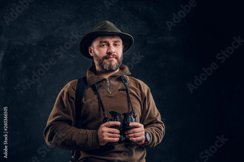 Mature hunter with rifle holding binoculars and looking at a camera. Studio photo against dark wall background