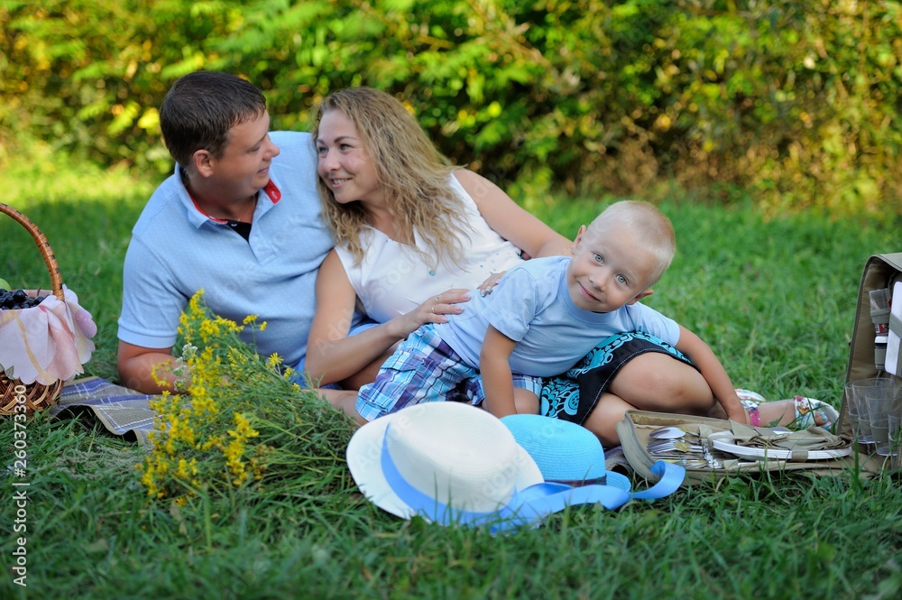 Fototapeta premium Family picnic in nature. Mom, dad and son relax sitting on the grass in the Park in the summer at sunset. A little boy looks at the camera and smiles. Portrait. Horizontal orientation of the image