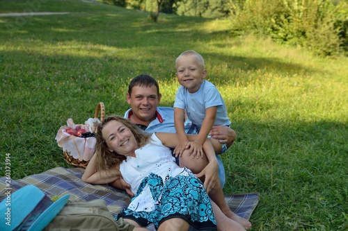Happy mom, dad and baby smile and look at the camera while lying on the blanket in the Park at sunset. Family picnic outdoors. Portrait. Horizontal orientation