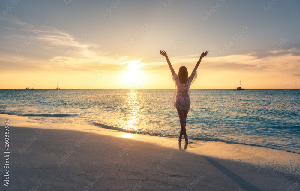 Beautiful young woman standing in sea with waves on sandy beach against orange sky at sunset. Summer travel. Happy slim girl in white lace dress with raised up arms on the seashore in Zanzibar, Africa