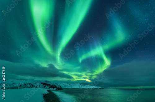 Aurora borealis over ocean. Northern lights in Teriberka  Russia. Starry sky with polar lights and clouds. Night winter landscape with aurora  sea with stones in blurred water  snowy mountains. Travel