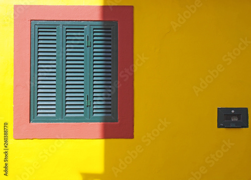 a vibrant yellow wall with red frame and green closed shuttered window typical Portuguese colors in bright sunlight and shade with space for copy