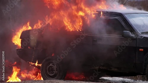Massive orange flame of fire with huge matt black smoke burns rusty dirty old dark dusty violet car which is left on deserted grey road at some suburb area on cold cloudy winter day.