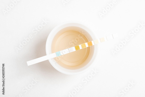 A set of urine sample on a round plastic cup with a single unused urine reagent strip against a plain white background.