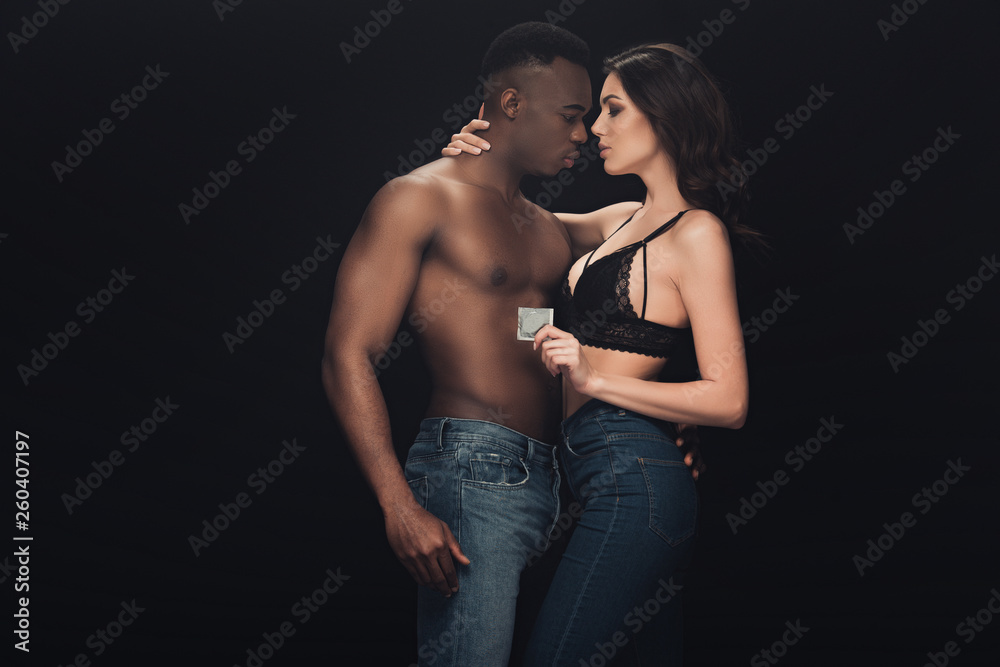 beautiful sexy woman embracing shirtless african american man and holding condom isolated on black beautiful sexy woman embracing shirtless african american man and holding condom isolated on black