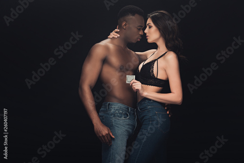 beautiful sexy woman embracing shirtless african american man and holding condom isolated on black