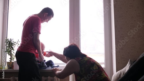 senior man and woman are playing with their adorable little grandson sitting on the window sill, and grandmother scaring the boy with her hands and the kid is smiling in slow motion middle shot 4K