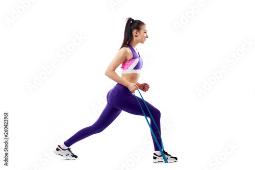 Sporty woman does exercises on legs, makes lunges with sport fitness rubber bands on white background. Photo of muscular woman in sportswear on white background. Strength and motivation.