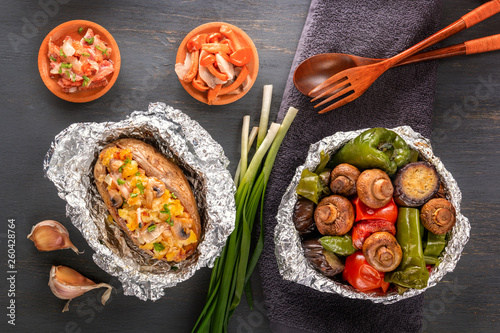 Baked potatoes with bacon, onions and baked vegetables in foil - tomatoes, eggplants, peppers on a gray wooden table. Top view