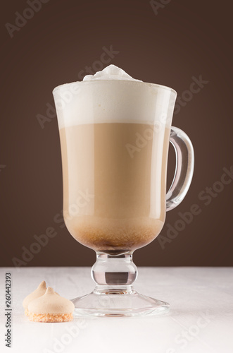 Cappuccino coffee with foam and cookies in transparent glass with handle on dark brown wall and white wood board.