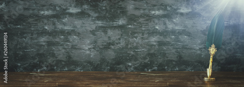 Old feather quill ink pen with inkwell over wooden desk infront of black wall background. Vintage old style and filtered. Conceptual photo on history, education, fantasy and literature topic