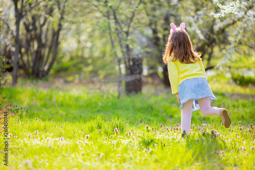 Adorable little girl sitting at the green grass playing in the garden on Easter egg hunt.