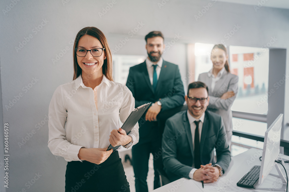 Smiling Caucasian businesswoman in formal wear, with eyeglasses and brown hair holding clipboard while standing in office. In background coworkers smiling and posing. - obrazy, fototapety, plakaty 