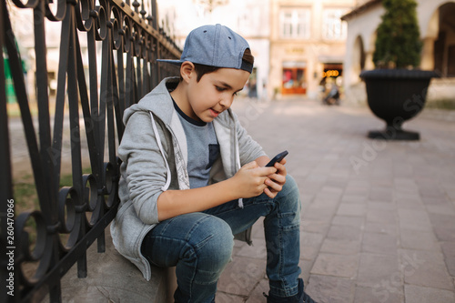 Young boy play online game at smartphone outside. Boy sitting in centre of the city. Boy play games while his mom going to the shop © Aleksandr