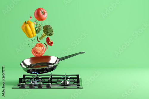Fresh colored vegetables and glass lid are flying from the frying pan. At the bottom - a cooking stove with burning fire. Green studio background