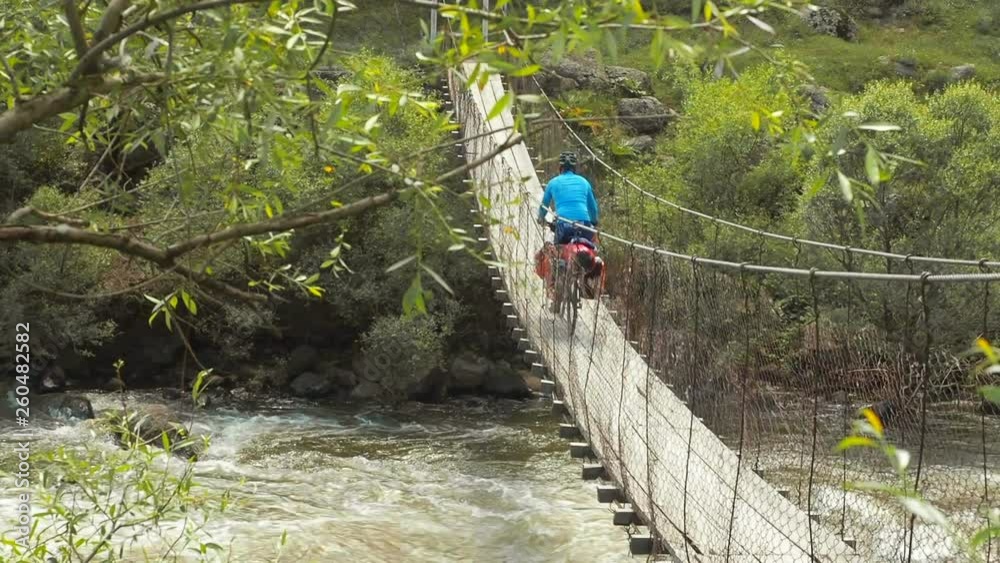 Bicyclist rides a suspension bridge stretched across a stormy mountain river. A cyclist rides on a bicycle along a long narrow wooden suspension bridge that stretches between the banks of a turbulent Bicyclist rides a suspension bridge stretched across a stormy mountain river. A cyclist rides on a bicycle along a long narrow wooden suspension bridge that stretches between the banks of a turbulent