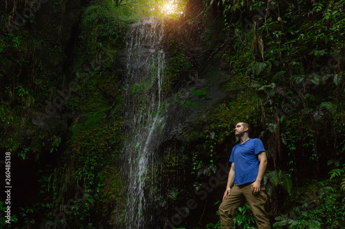 A brave explorer stands in the middle of the dark jungle near the waterfall and looks up at the ray of sunshine. travel without life insurance